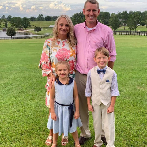 A family of four poses outdoors on a grassy field. The woman wears a floral dress, the man a pink shirt, and the children are in light-colored outfits. A pond and trees are visible in the background under a partly cloudy sky.