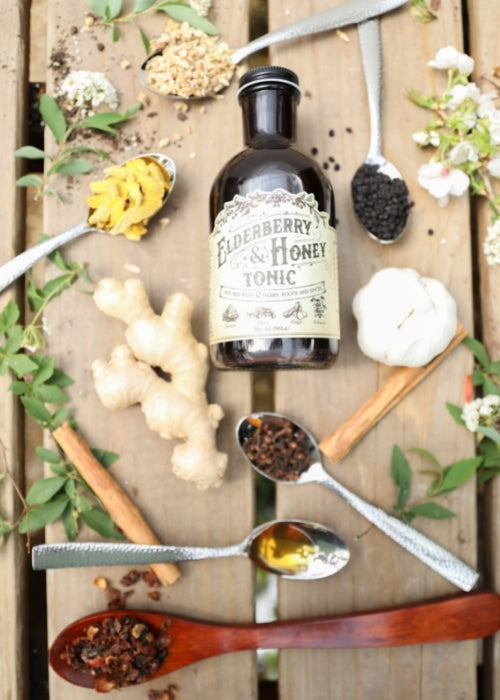 Elderberry and honey tonic by Roots and Leaves LLC in a glass bottle, with fresh elderberries and honey drizzled around it.