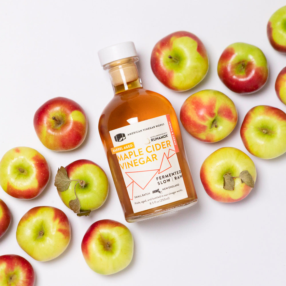 Bottle of American Vinegar Works Maple Cider Vinegar on a kitchen counter with fresh ingredients and a bowl of salad.