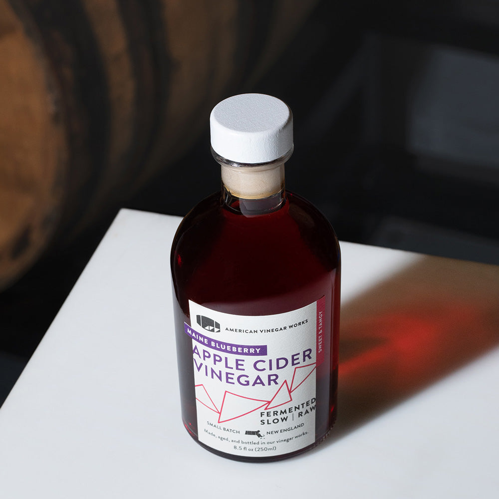 Bottle of American Vinegar Works wild blueberry apple cider vinegar with blueberries and apples in a lifestyle setting.