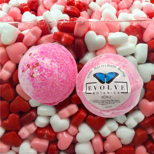 Evolve Botanica XOXO bath bomb with heart-shaped design, vibrant pink and purple colors, floating in a clear bathwater.