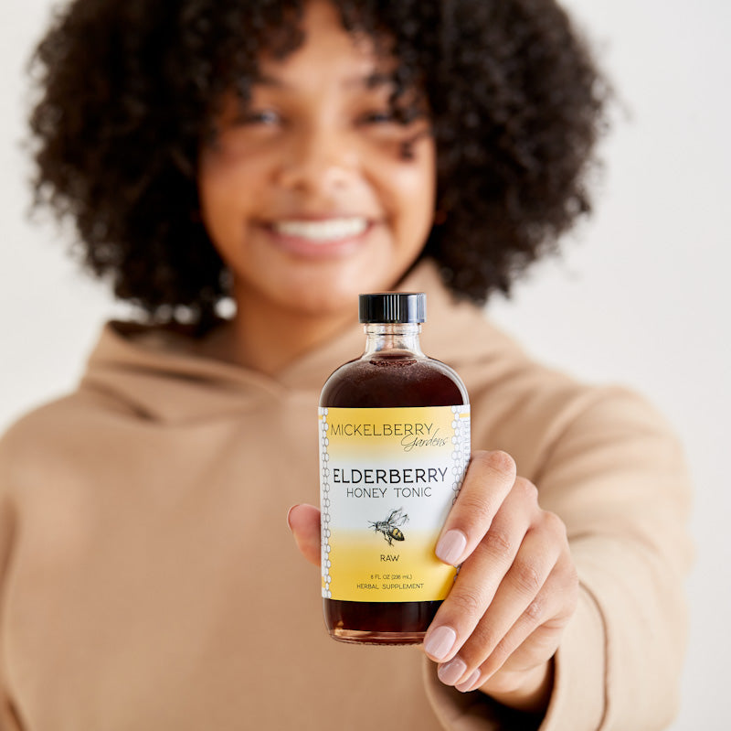 Mickelberry Gardens Elderberry Honey Tonic in an 8oz bottle, held by a model, featuring dark elderberry syrup with honey.