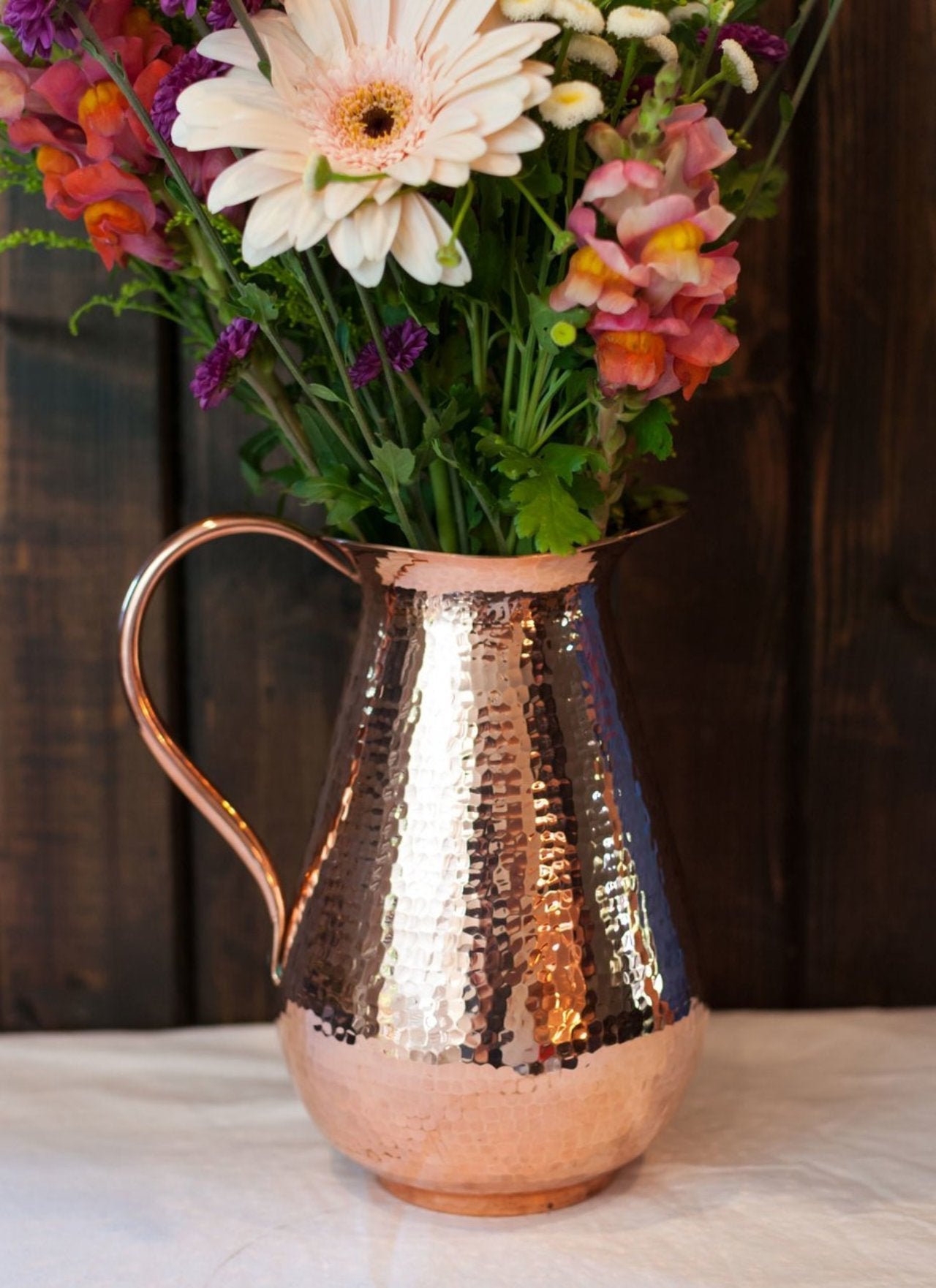 Sertodo Bisotun copper water pitcher with a curved handle, resting on a wooden surface in a natural setting.