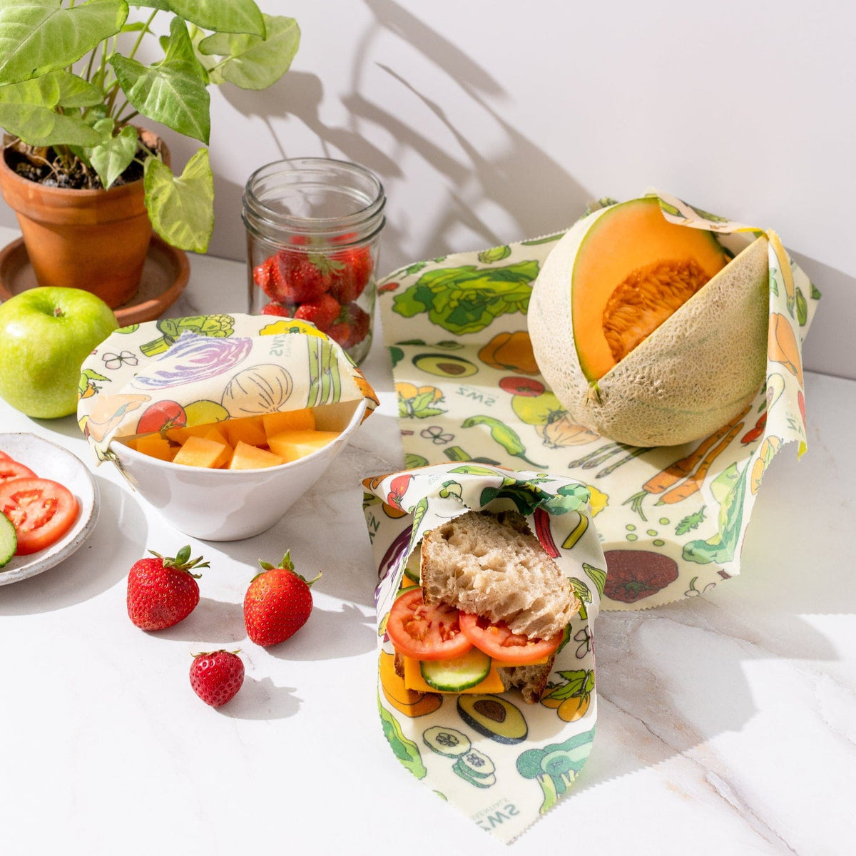 Beeswax wraps covering veggies, a sandwich, and a melon, showcasing plastic-free food storage by ZeroWasteStore.com.