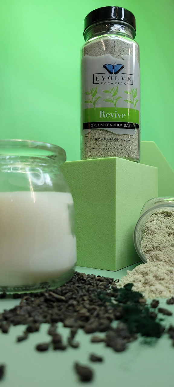 Evolve Botanica Milk Bath Revive with Green Tea in a glass jar, creamy powder with green tea leaves visible inside.