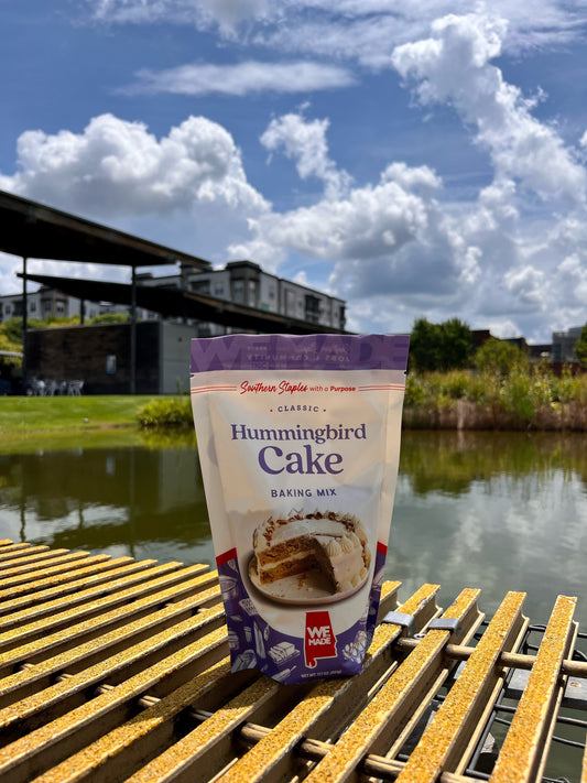 A box of WE Made Hummingbird Cake Mix with vibrant packaging, featuring images of a freshly baked hummingbird cake.